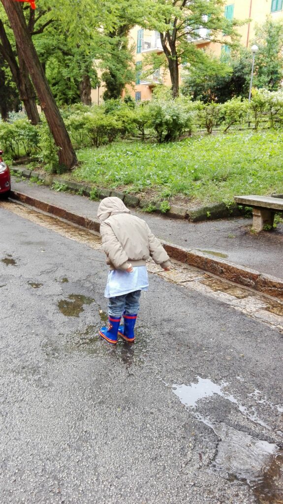 비오는날 장화를 신고 유치원 가운을 입은 아이가 물웅덩이에서 폴짝인다. 주변에는 나무들과 풀이 보인다