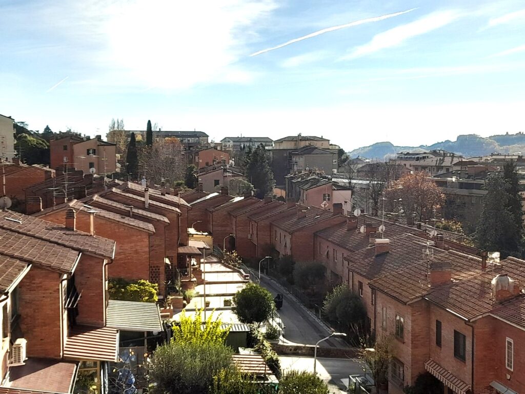 붉은 벽돌 지붕이 이어진 이탈리아 주택가와 골목길, 맑은 하늘 아래 멀리 보이는 언덕 풍경