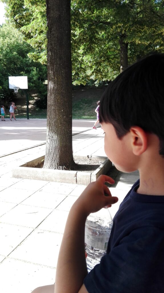 나무와 농구경기장이 있는 공원의 벤치에 앉아있는 아이의 옆모습