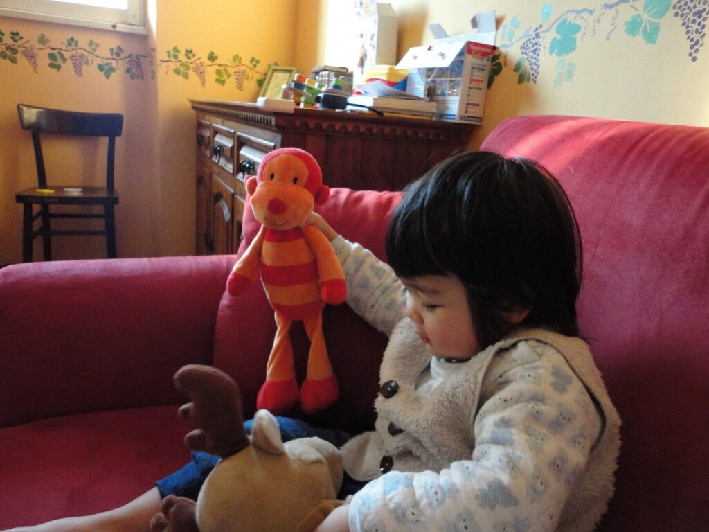 벽이 노란 집에서 어린 남자 아이가 빨간색과 주황색 스트라이프 무늬의 원숭이 인형을 가지고 빨간색 소파 위에서 놀고 있다.