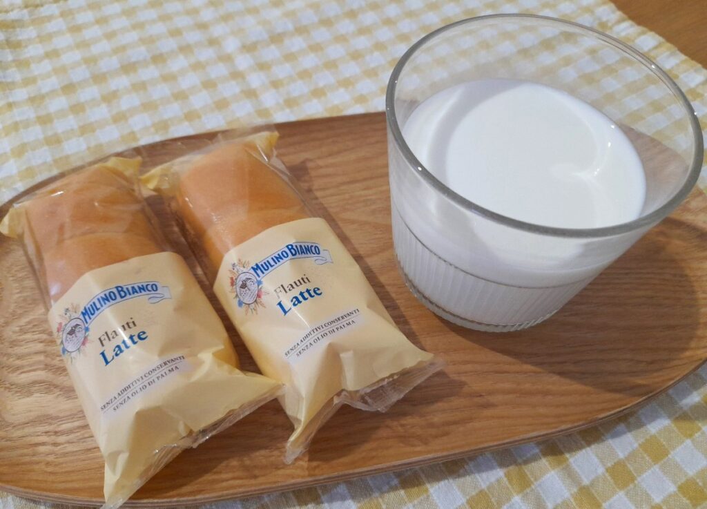 나무 트레이 위에 Mulino Bianco Flauti Latte 빵 두 개와 우유 한 잔이 놓여 있다