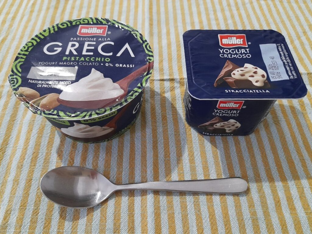 테이블매트 위에 뮐러 그릭 피스타치오 요거트와 스트라치아텔라 요거트, 스푼이 함께 놓여 있다