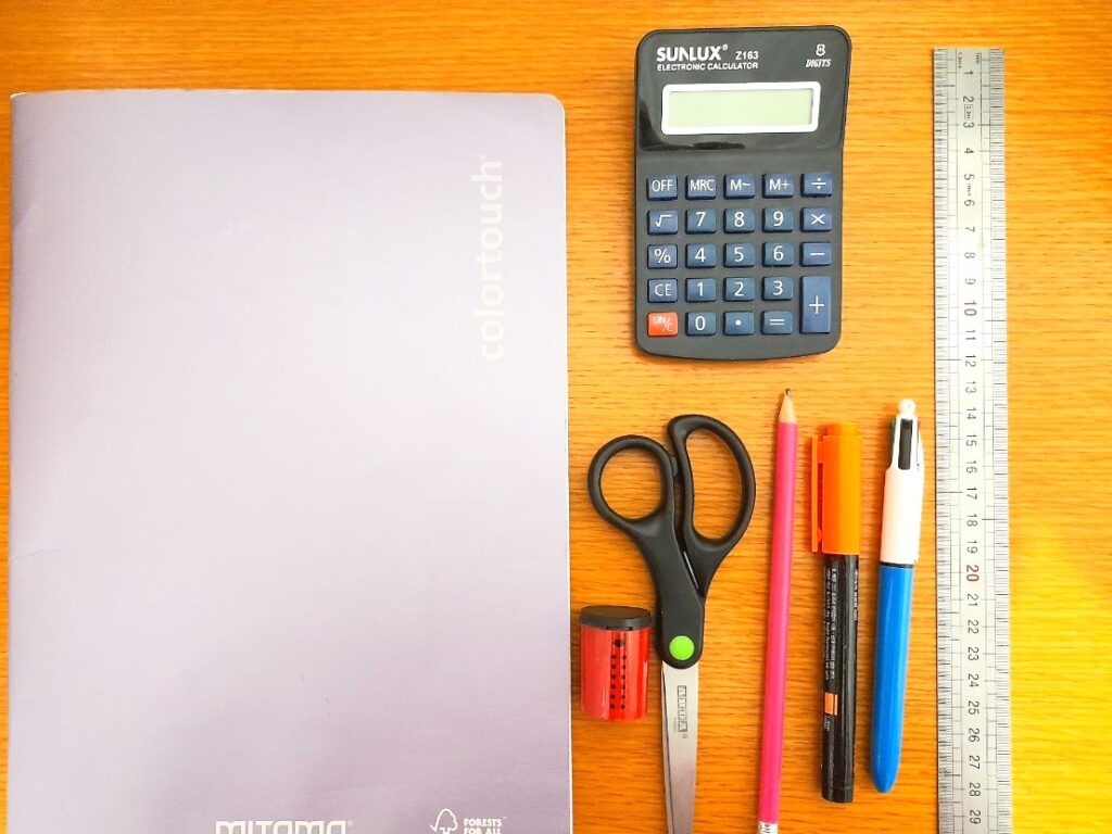 나무 책상 위에 공책과 계산기, 자, 가위, 연필과 펜 등 학용품이 가지런히 놓여 있다