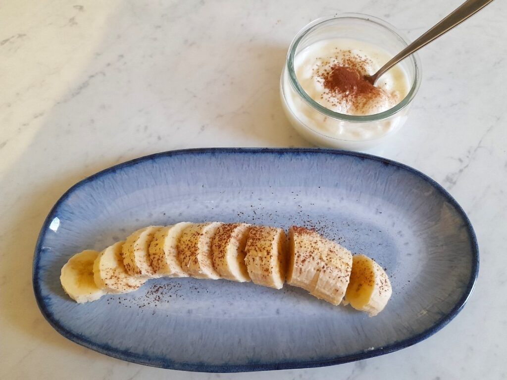 접시에 바나나를 썰어 담고 시나몬을 뿌렸으며, 옆에 시나몬을 올린 요거트 한 컵이 놓여 있다